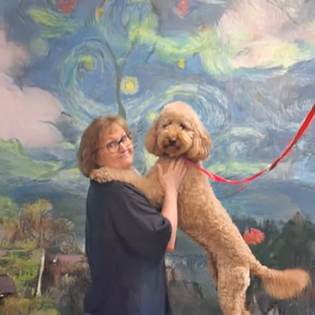 Smiling groomer holds a fluffy Goldendoodle on a red leash in front of a Van Gogh-style mural at Empire Pets.