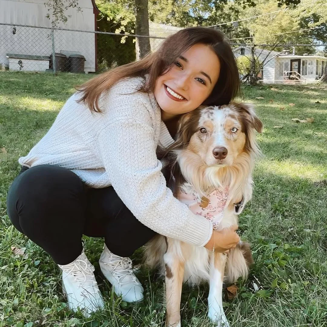 Smiling woman in a sweater hugs a red merle Australian Shepherd wearing a pink harness while posing on the grass.